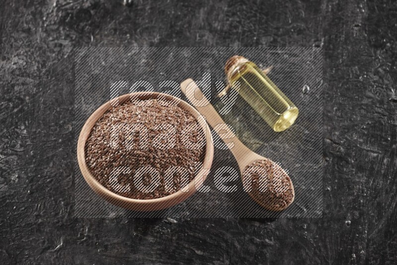 A wooden bowl and spoon full of flaxseeds and a glass bottle of flaxseeds oil on a textured black flooring