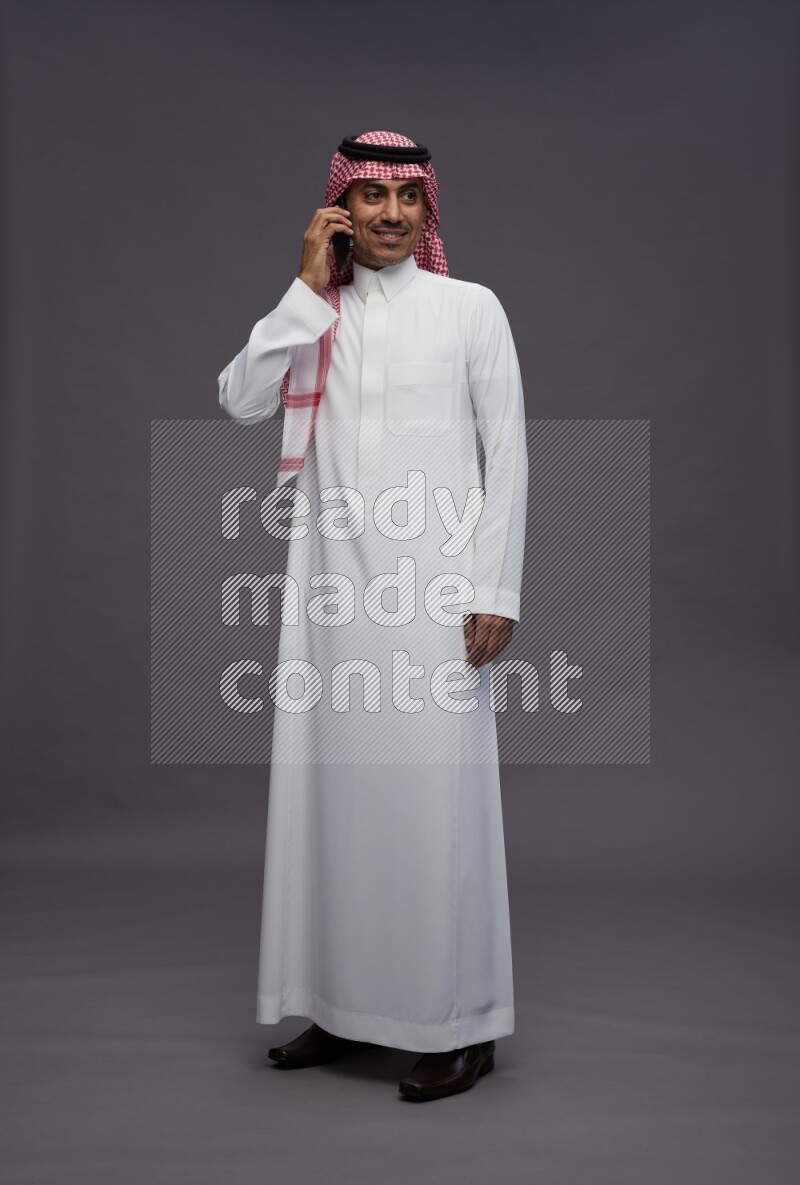 Saudi man wearing thob and shomag standing talking on phone on gray background