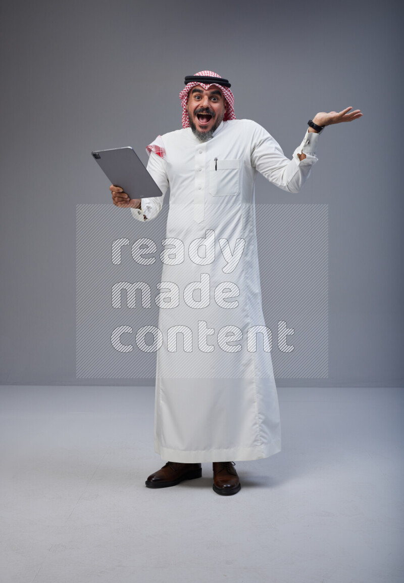 Saudi man Wearing Thob and red Shomag standing working on tablet on Gray background