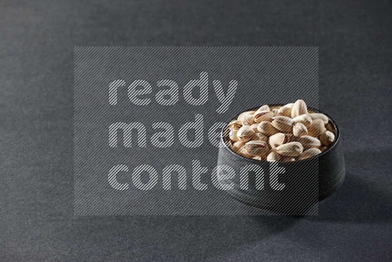 A black pottery bowl full of pistachios on a black background in different angles