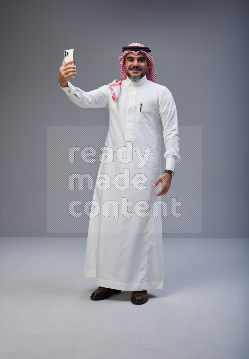 Saudi man Wearing Thob and red Shomag standing taking selfie on Gray background