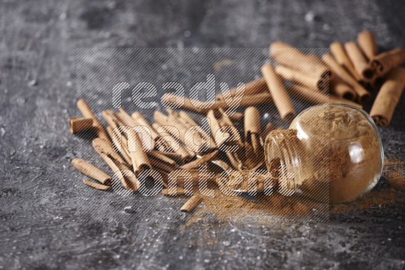 Herbal glass jar full cinnamon powder flipped and a metal spoon full of powder surrounded by cinnamon sticks on textured black background in different angles