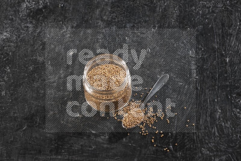 A glass jar and a metal spoon full of mustard seeds on a textured black flooring