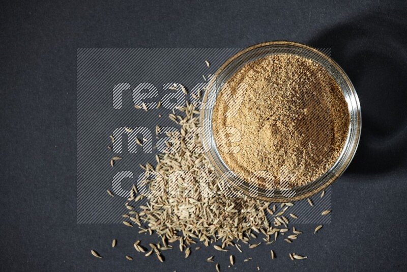 A glass bowl full of cumin powder with cumin seeds beside it on black flooring
