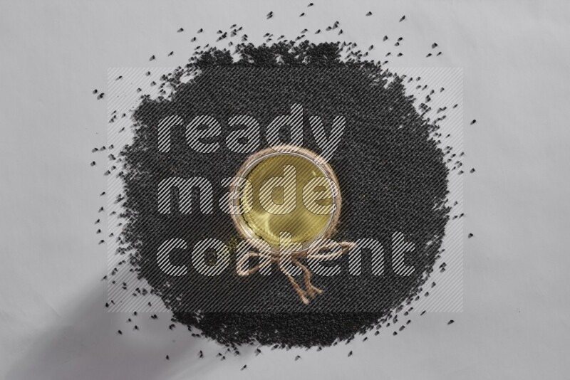 A glass jar full of black seeds oil surrounded by the seeds on a white flooring