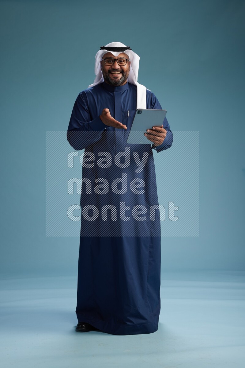 Saudi Man with shimag Standing holding ATM card while working on tablet on blue background