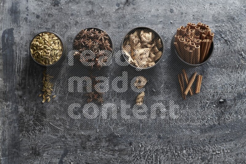 Ginger, Cardamom, Star anise and cinnamon sticks in 4 bowls on a textured black background