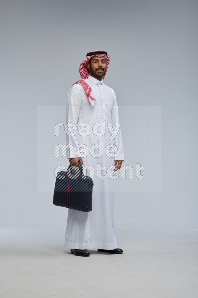 Saudi man Wearing Thob and shomag standing holding bag on Gray background