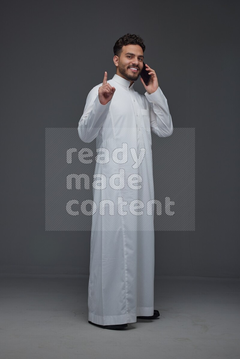 A Saudi man wearing Thobe and talking in the phone while standing and making different hand gestures eye level on a gray background