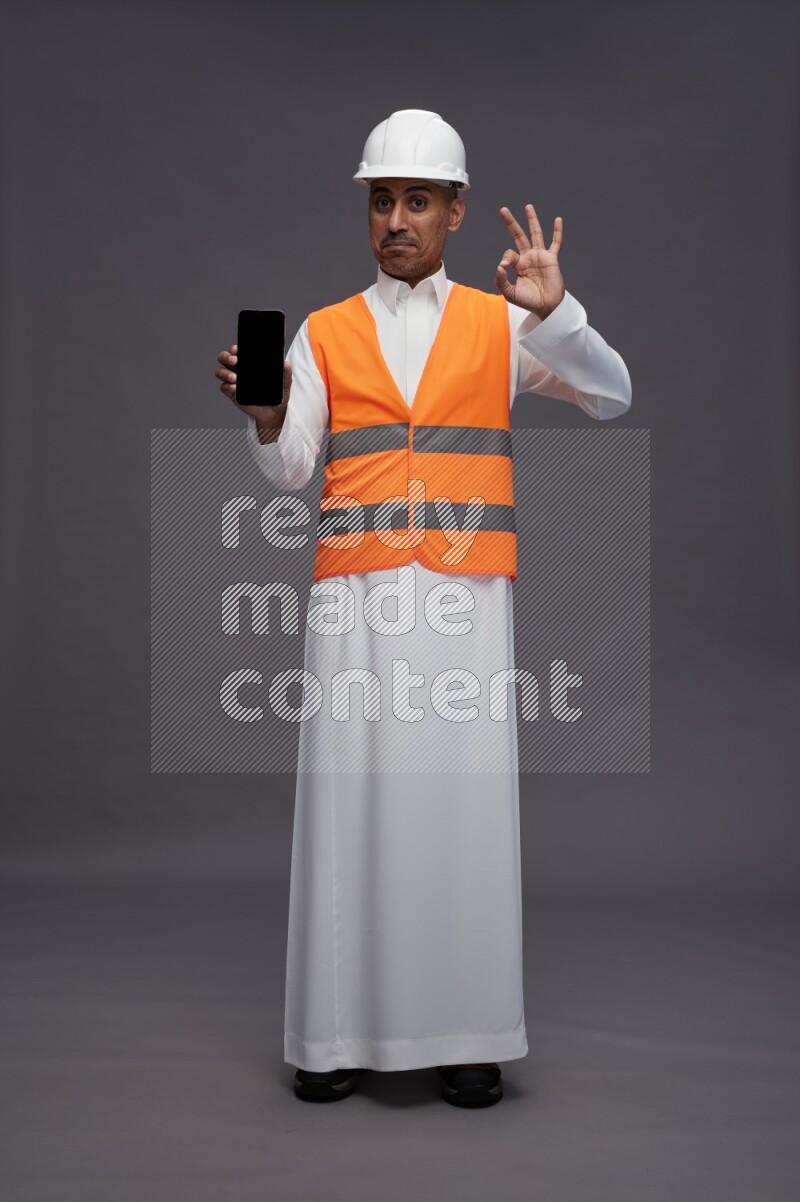 Saudi man wearing thob with engineer vest standing showing phone to camera on gray background