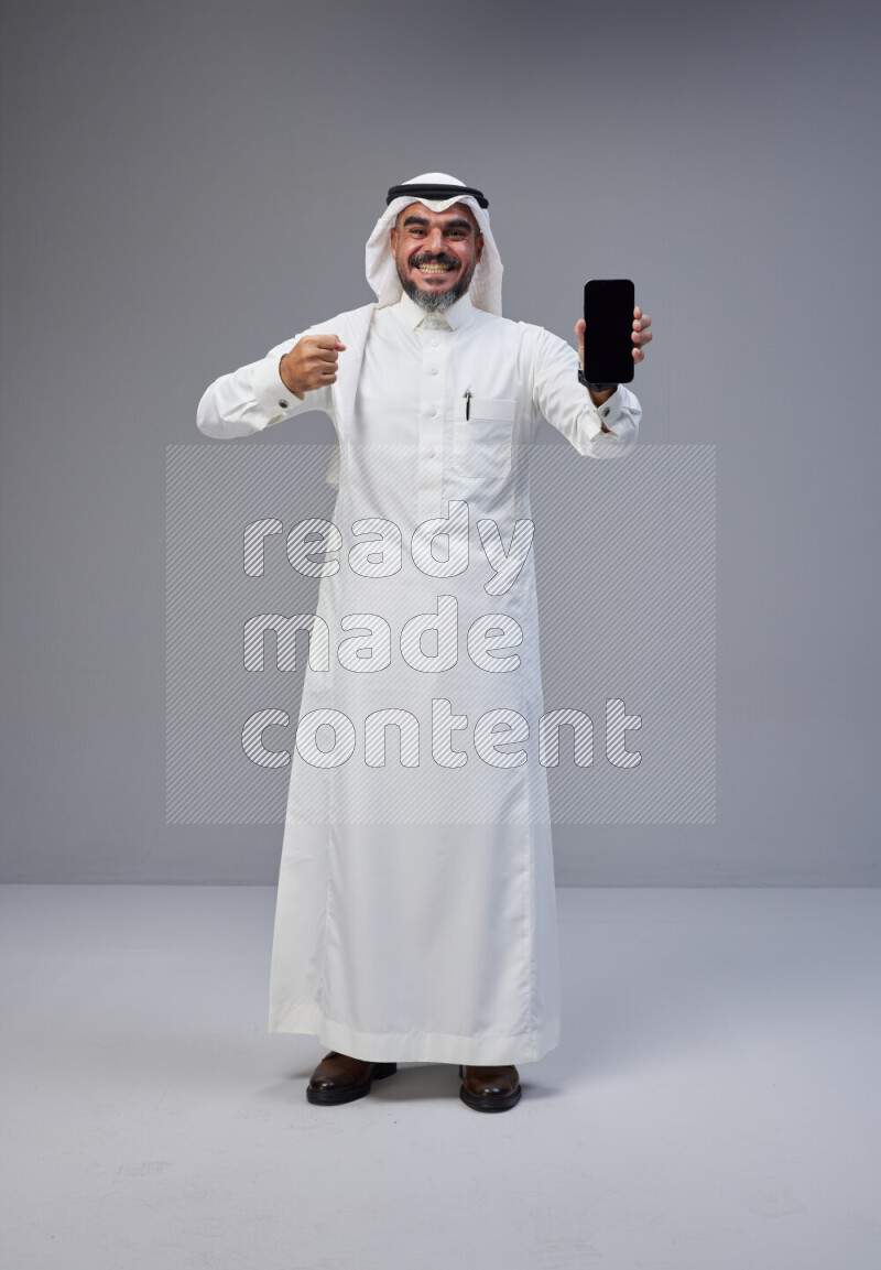 Saudi man Wearing Thob and white Shomag standing showing phone to camera on Gray background