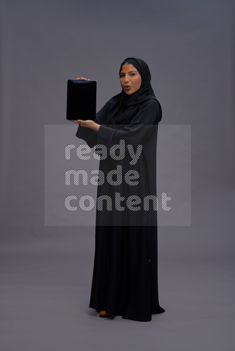 Saudi woman wearing Abaya standing showing tablet to camera on gray background