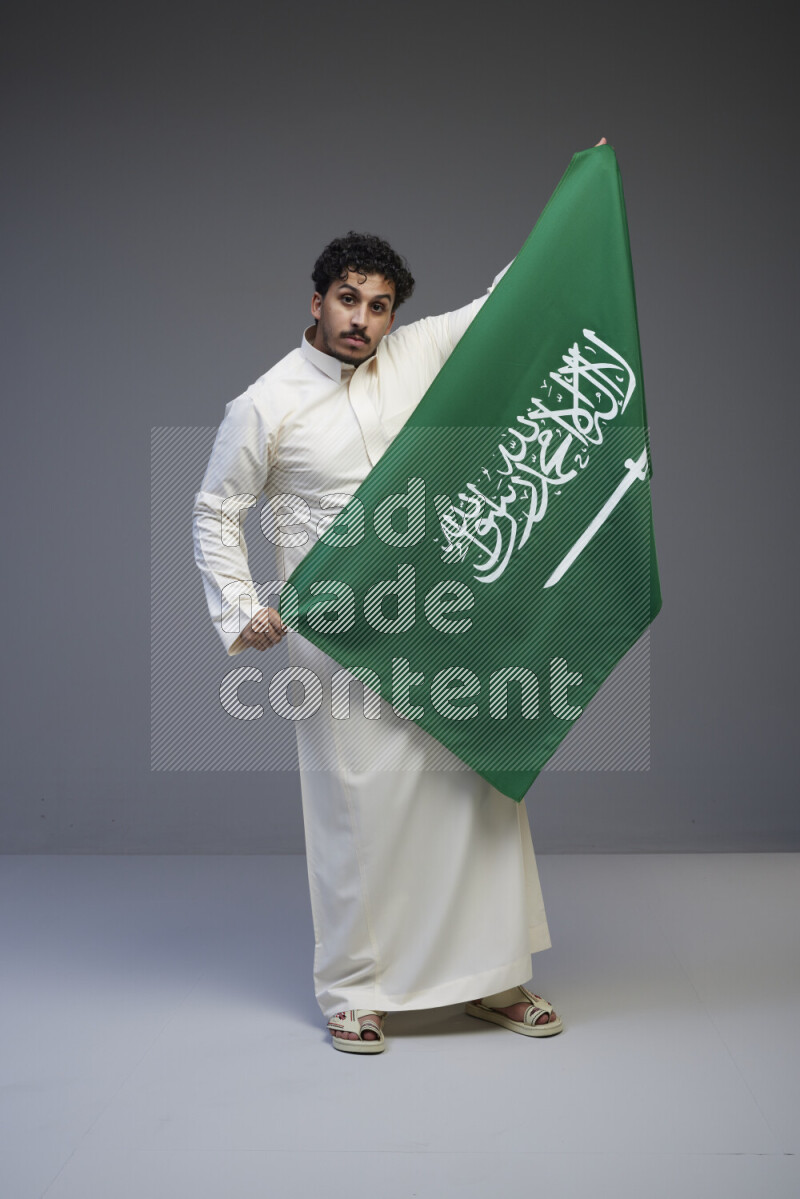 A saudi man standing wearing thob holding big saudi flag on gray background