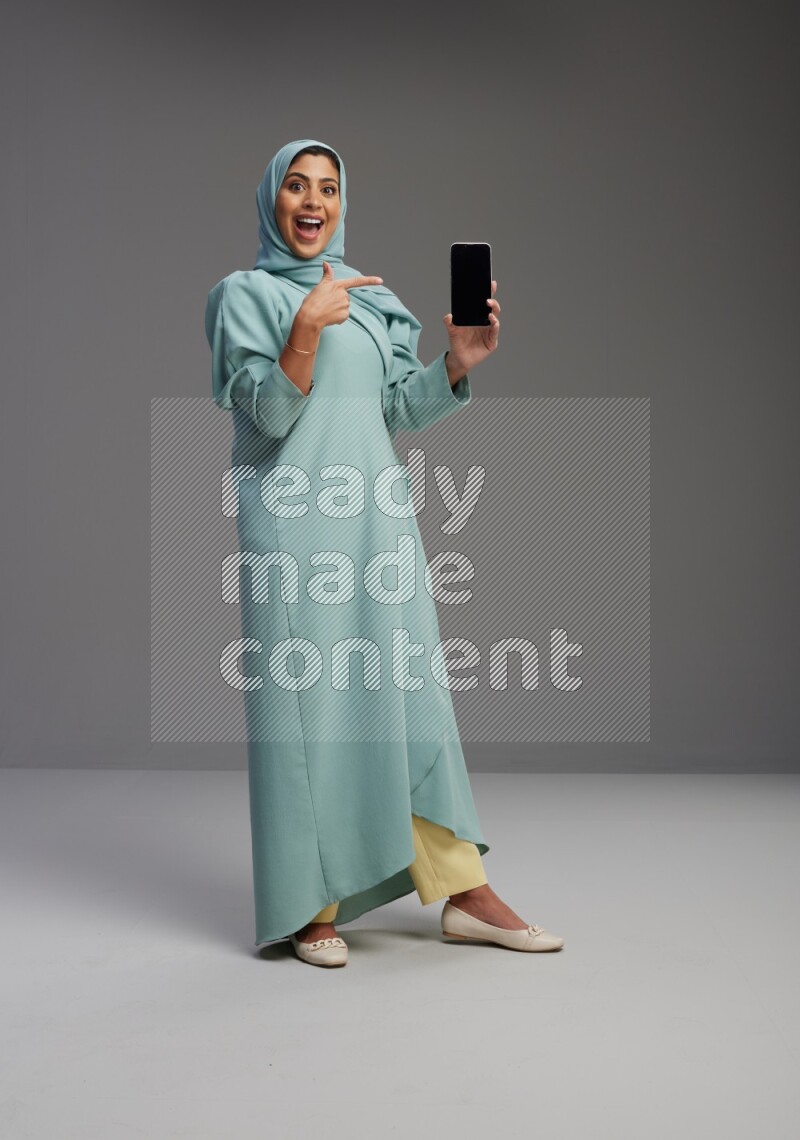 Saudi Woman wearing Abaya standing showing phone to camera on Gray background