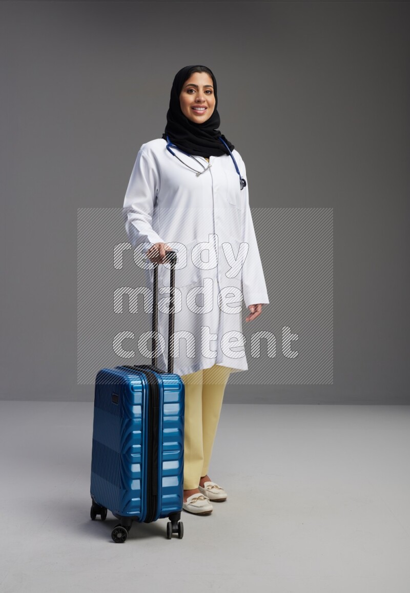 Saudi woman wearing lab coat with stethoscope standing holding Travel bag on Gray background