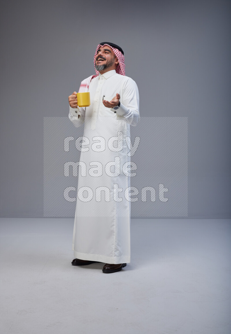 Saudi man Wearing Thob and red Shomag standing holding mug on Gray background