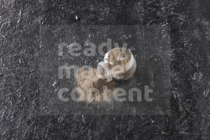 A flipped glass spice jar full of black pepper powder on textured black flooring