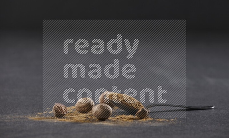 ملعقة معدنية ممتلئة ببودرة جوزة الطيب مع بودرة وحبوب جوزة الطيب بجانبها على خلفية سوداء