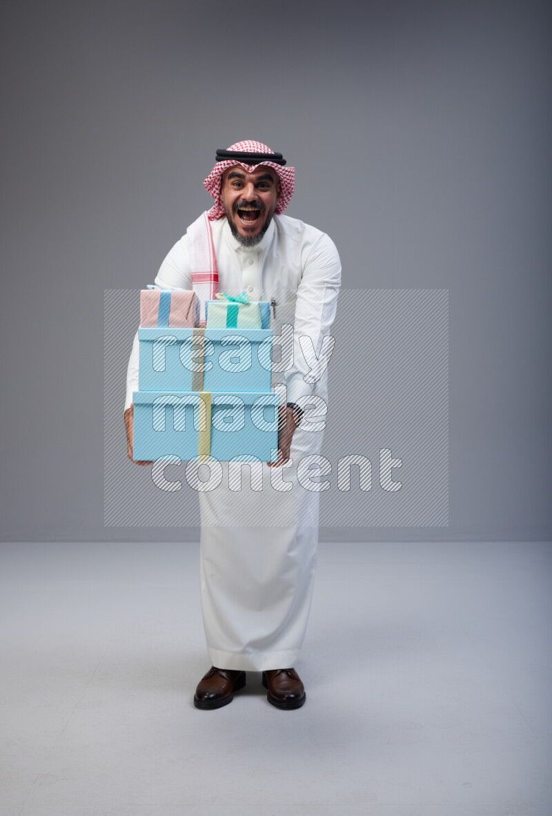 Saudi man Wearing Thob and red Shomag standing holding gift box on Gray background