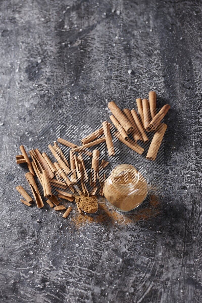 Herbal glass jar and a metal spoon full of cinnamon powder surrounded by cinnamon sticks on textured black background