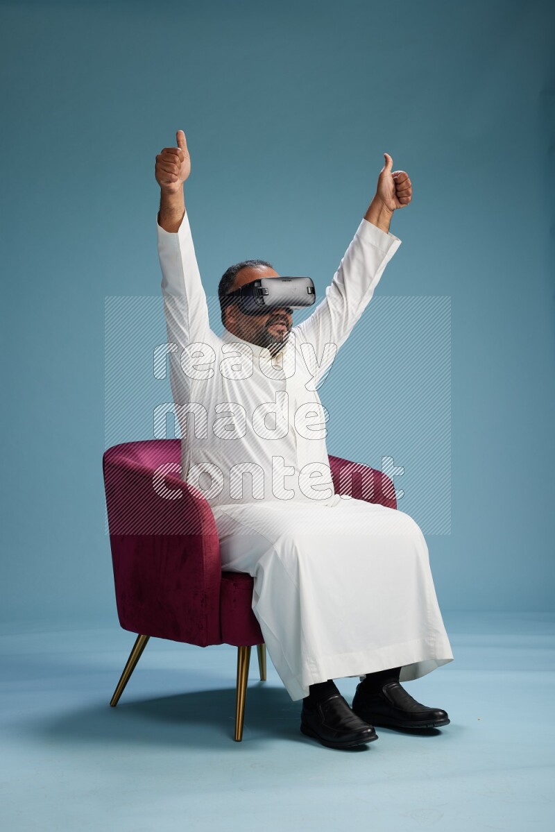 Saudi Man without shimag sitting on chair  gaming with VR on blue background