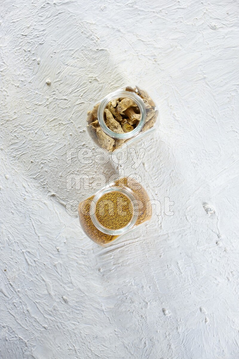 وعاءان زجاجيان للتوابل ممتلئان ببودرة الكركم وأصابع الكركم المجففة على خلفية بيضاء