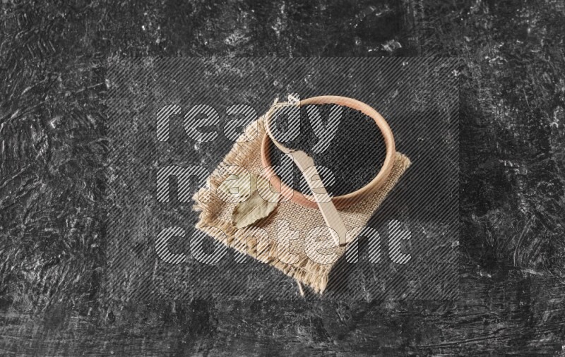 A wooden bowl full of black seeds with wooden spoon full of the seeds on it on a burlap fabric on a textured black flooring