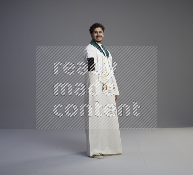 A Saudi man standing wearing thob and Saudi flag scarf showing phone to camera on gray background