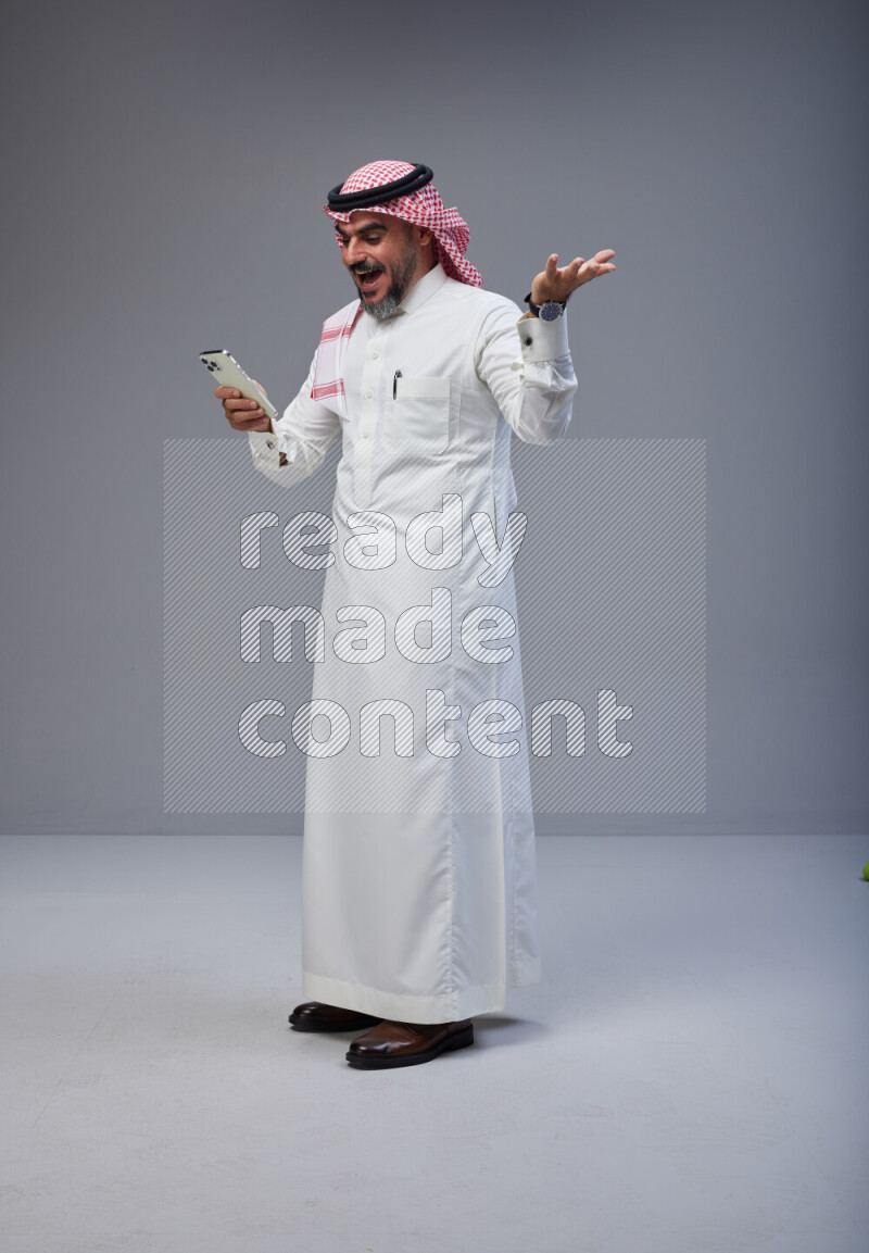 Saudi man Wearing Thob and red Shomag standing texting on phone on Gray background