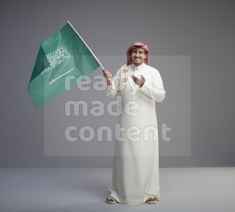 A Saudi man standing wearing thob and red shomag raising big Saudi flag on gray background