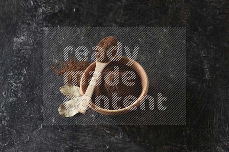 A wooden bowl and a wooden spoon full of cloves powder with laurel leaves on a textured black flooring