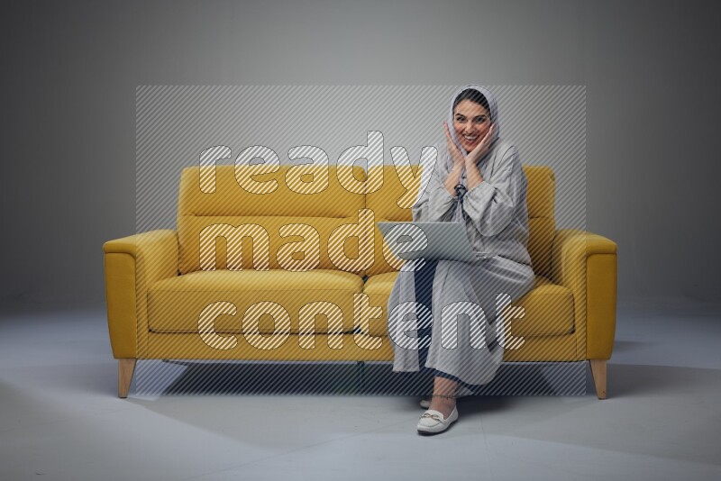A Saudi woman wearing a light gray Abaya and head scarf sitting on a yellow sofa and using her laptop eye level on a grey background