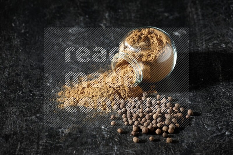 A flipped glass spice jar full of allspice powder and powder spilled out of it with whole balls on a textured black flooring
