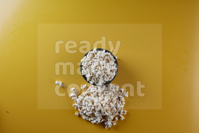 A black ceramic bowl full of popcorn with popcorn beside it on a yellow background in different angles