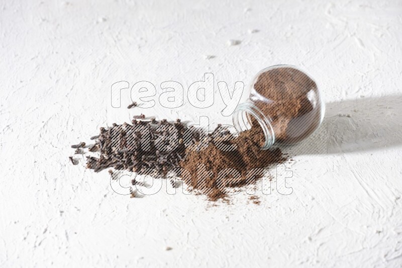 A glass spice jar full of cloves powder flipped and powder came out of it with cloves spread on textured white flooring