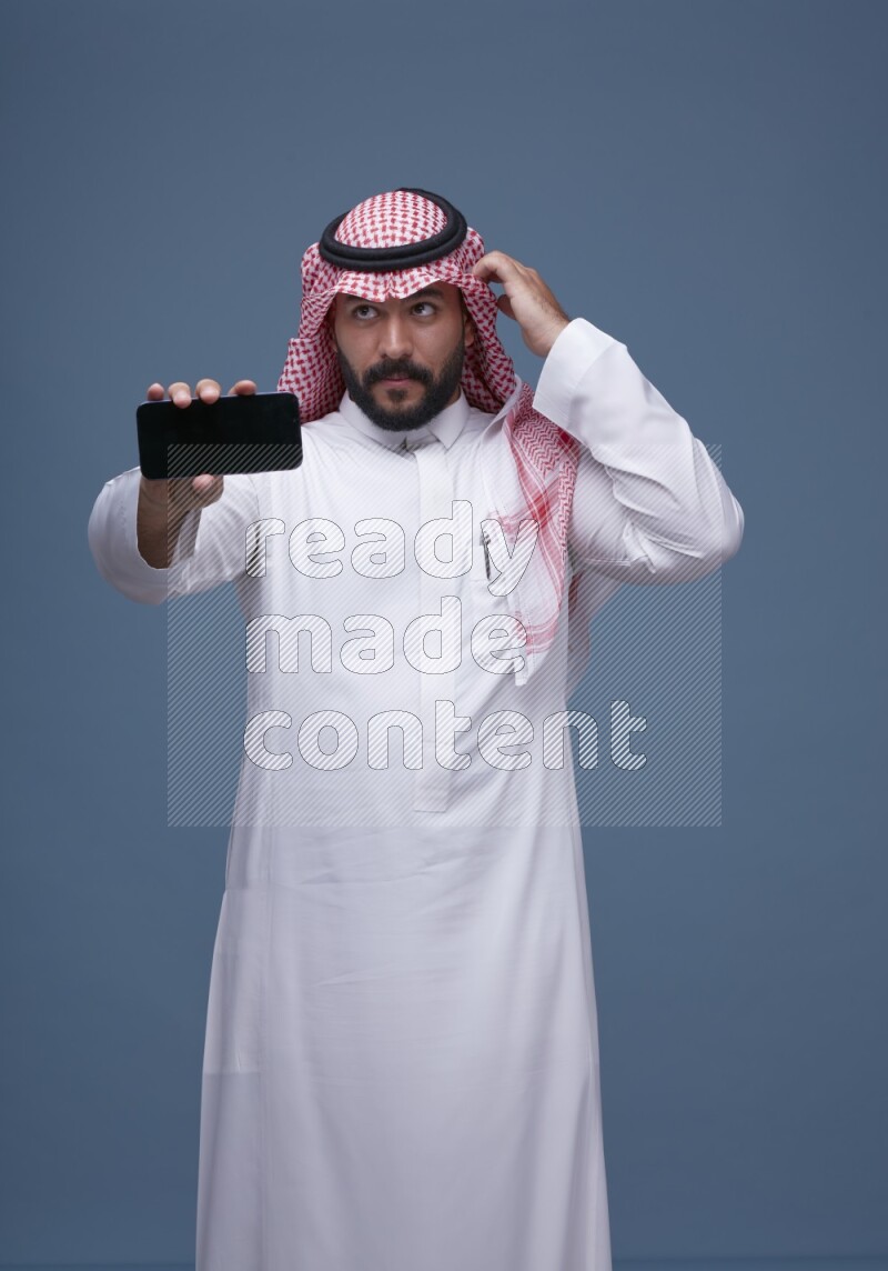 A man Showing his smartphone Screen on Blue Background wearing Saudi Thob