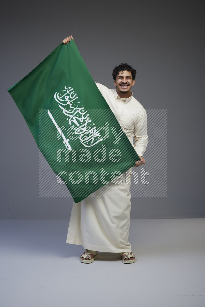 A Saudi man standing wearing thob holding big Saudi flag on gray background