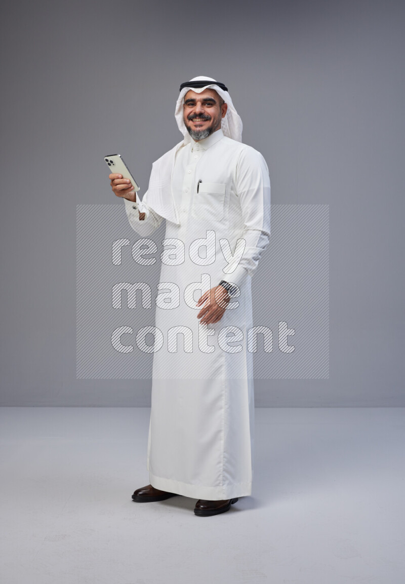 Saudi man Wearing Thob and white Shomag standing texting on phone on Gray background