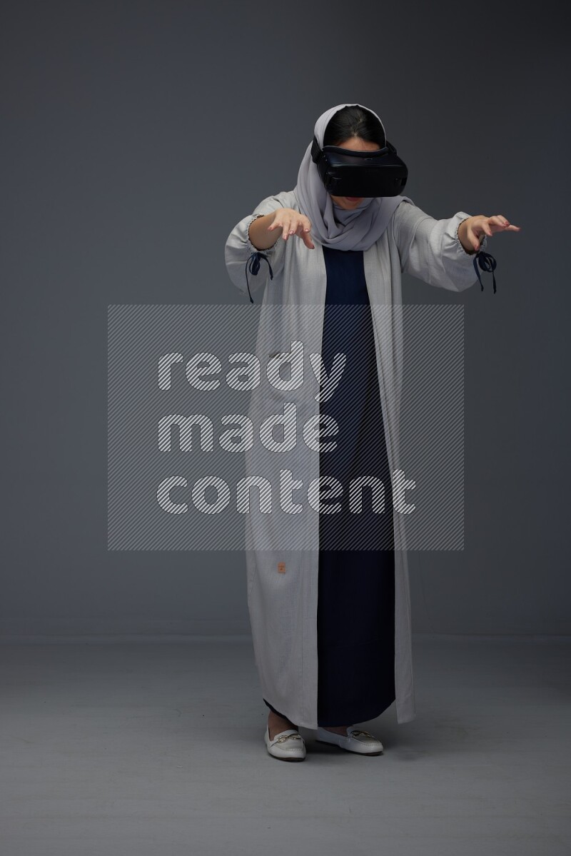 A Saudi woman wearing a light gray Abaya and head scarf standing and wearing VR eye level on a grey background