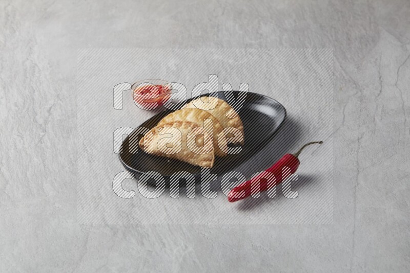 Three fried sambosas in an oval shaped black plate and a red sauce in a black round ramekin on a gray background