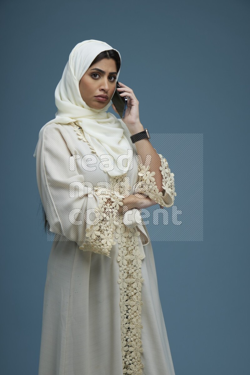 A Saudi woman having a call in a blue background wearing an off-white Abaya Hijab