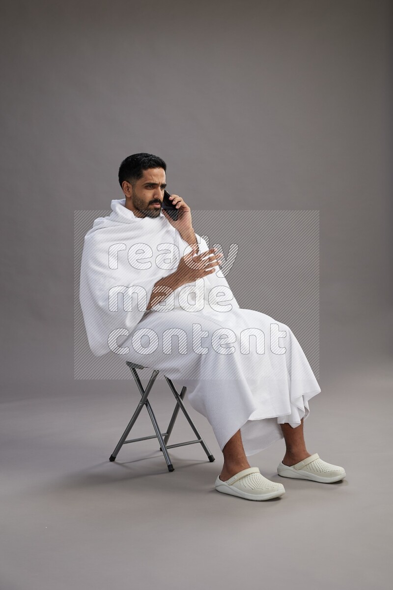 A man wearing Ehram sitting on chair talking on the phone on gray background