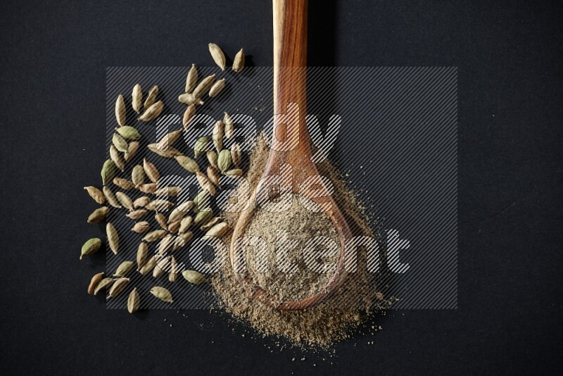 A wooden ladle full of cardamom powder and cardamom seeds beside it on black flooring