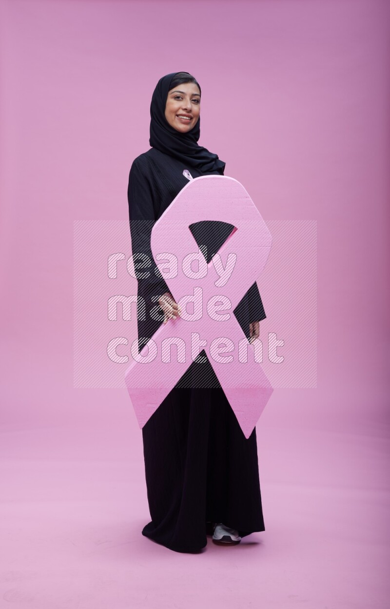 Saudi woman wearing pink ribbon on abaya standing holding awareness ribbon on plain pink background