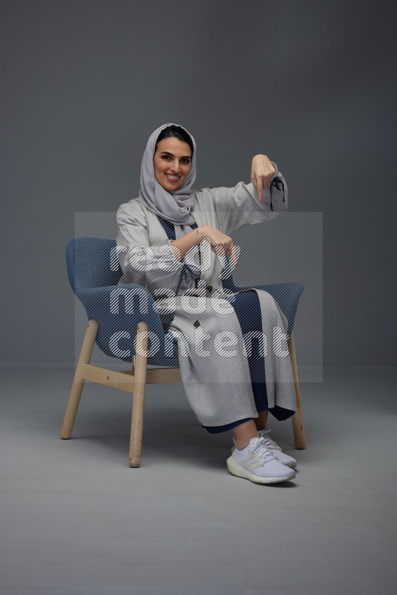 A Saudi woman wearing a light gray Abaya and head scarf sitting on a dark grey chair while making multi hand gestures eye level on a grey background