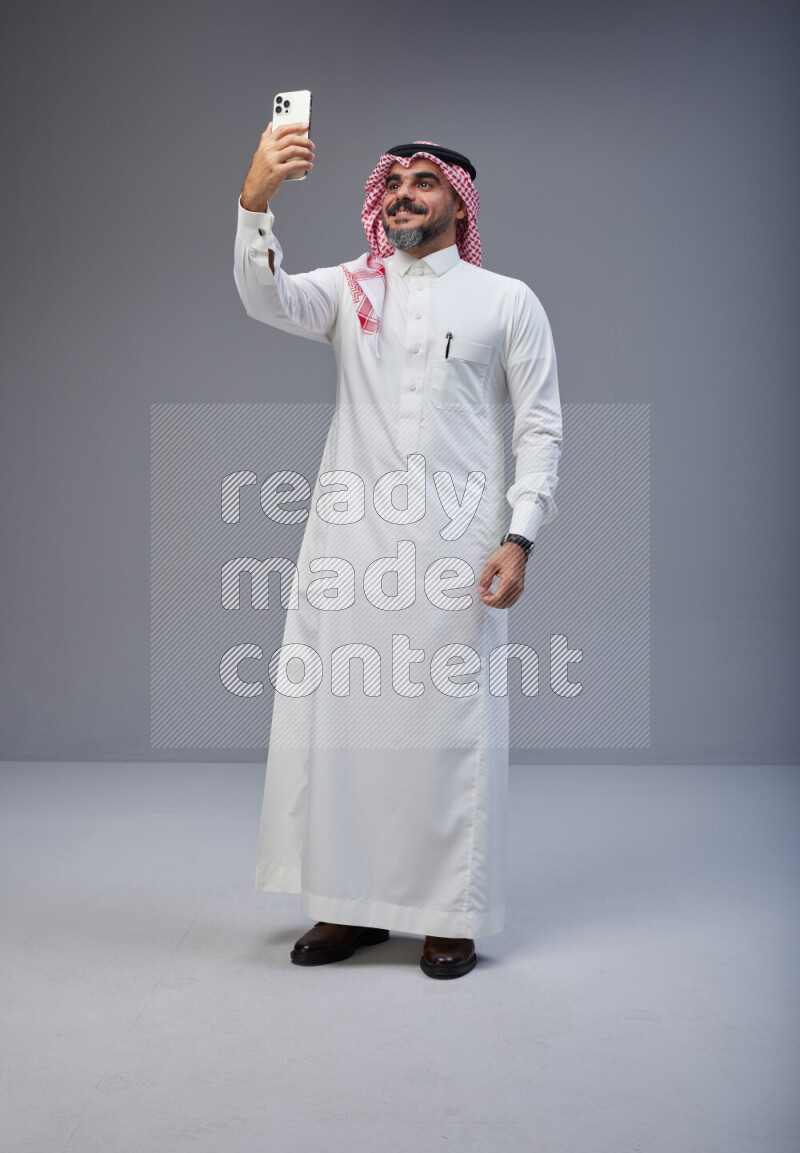 Saudi man Wearing Thob and red Shomag standing taking selfie on Gray background