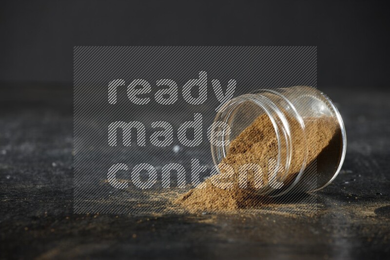 A flipped glass jar full of cumin powder and powder fell out on a textured black flooring