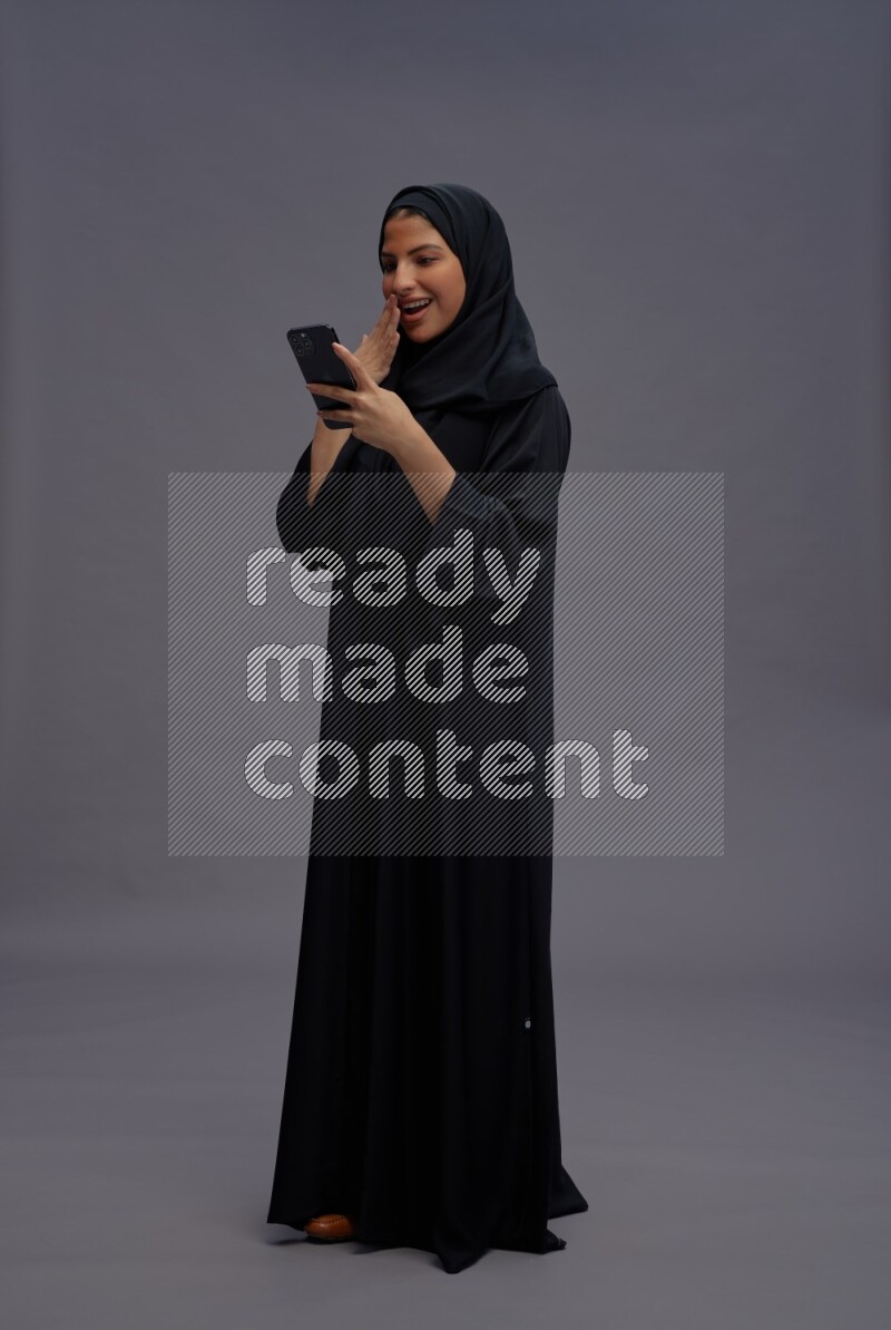 Saudi woman wearing Abaya standing texting on phone on gray background