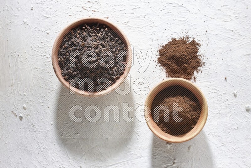 2 wooden bowls full of cloves powder and whole cloves on a textured white flooring