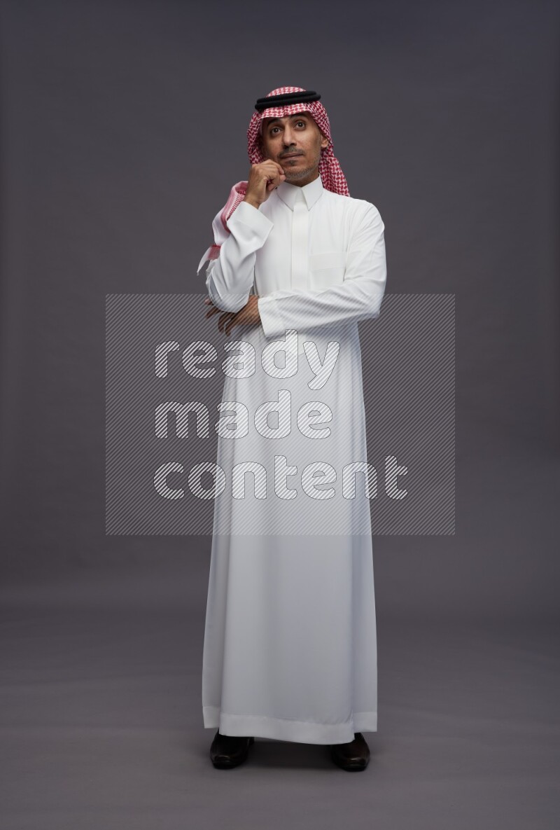 Saudi man wearing thob and shomag standing interacting with the camera on gray background